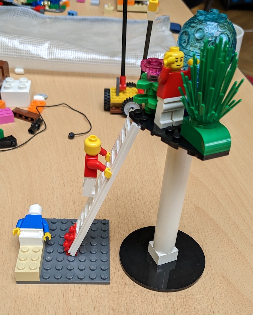 Lego model with one minifigure in a tree, another climbing a ladder into the tree and a third laying face down on the floor